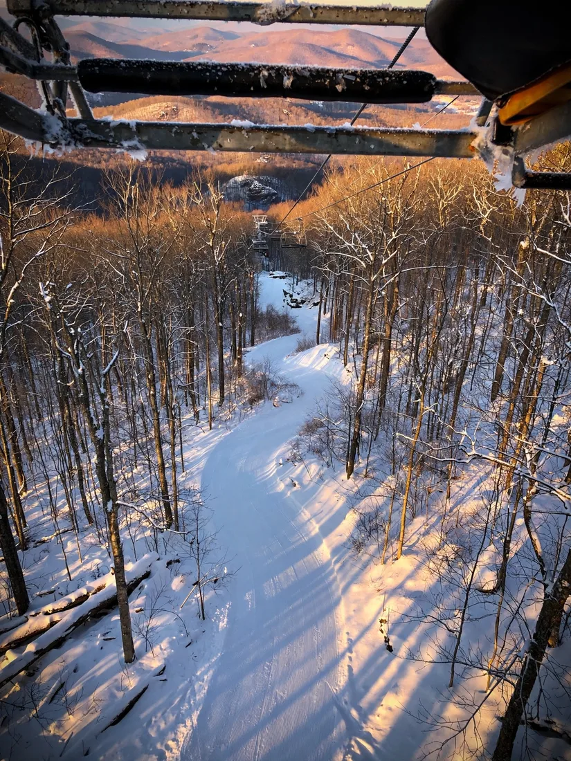 View from a chairlift over an empty groomed ski trail winding through snowy hardwood forest at golden hour